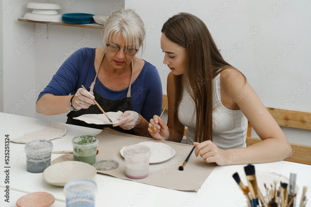 Woman potter with paintbrush glazing, painting on plate in workshop, working in pottery studio. People enjoying creative process, decorating ceramic after firing in oven. Hobby, small business Woman potter with paintbrush glazing, painting on plate in workshop, working in pottery studio. People enjoying creative process, decorating ceramic after firing in oven. Hobby, small business