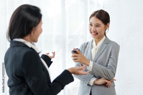 Two Asian business women discuss work with a smile while holding a cup of coffee in the office.