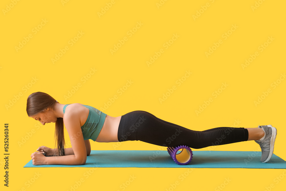 Young woman exercising with purple foam roller on yellow background