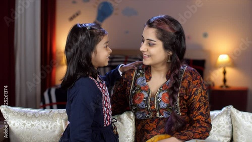 A little schoolgirl is whispering into her mother's ears - sharing her secrets  family bonding  mother-daughter love. An affectionate Indian mother and a young kid together at home - togetherness a... photo