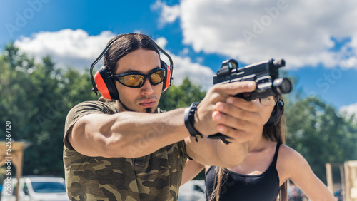 Dynamic closeup portrait of a handsome caucasian man in moro t-shirt and protective gear holding a black gun and shooting at the target. Beautiful sunny weather. Gun range concept. High quality photo