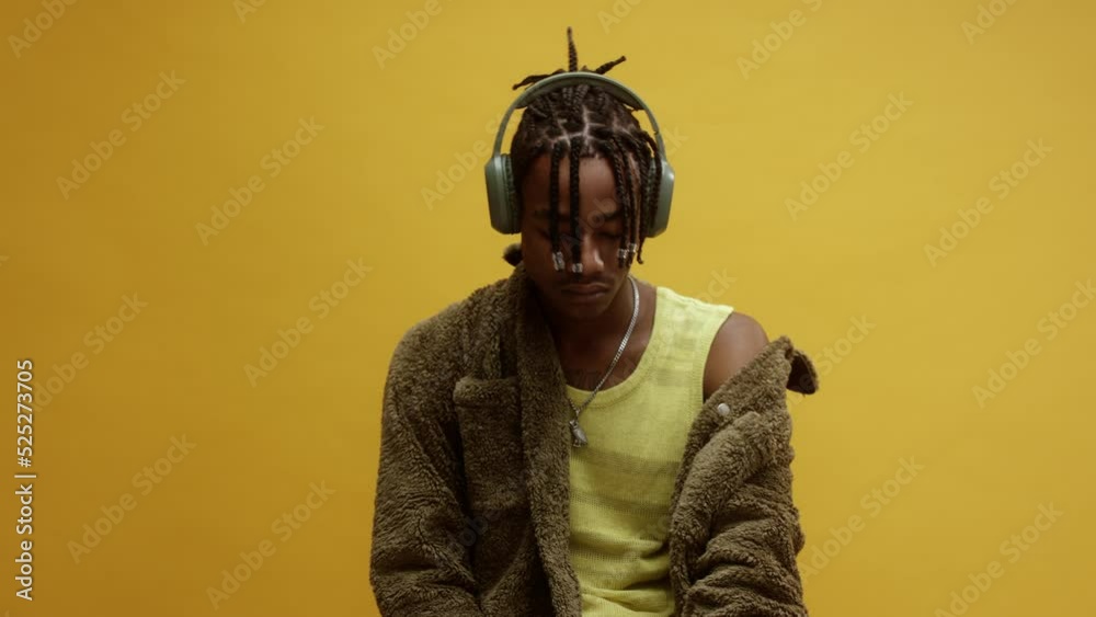 A young stylish black man, wearing a vest and white trousers, sitting on a chair enjoying some music on his headphones, in front of a yellow background