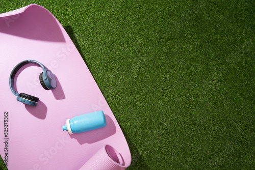 Yoga mat with headphones and water bottle on green grass. Sport gear.