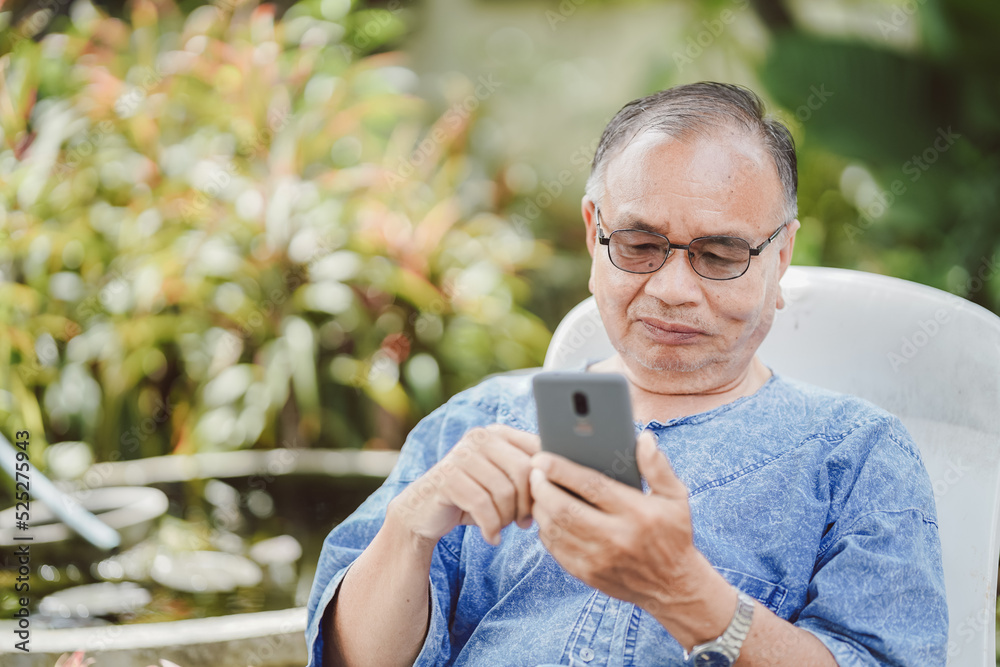 Fototapeta premium An old man is using a smartphone on a chair in the garden. Social network concept.