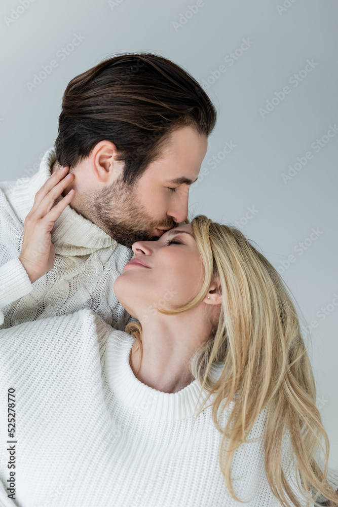 bearded man kissing forehead of blonde girlfriend in sweater isolated on grey