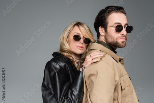 stylish blonde woman in sunglasses and leather jacket leaning on shoulders on bearded man isolated on grey