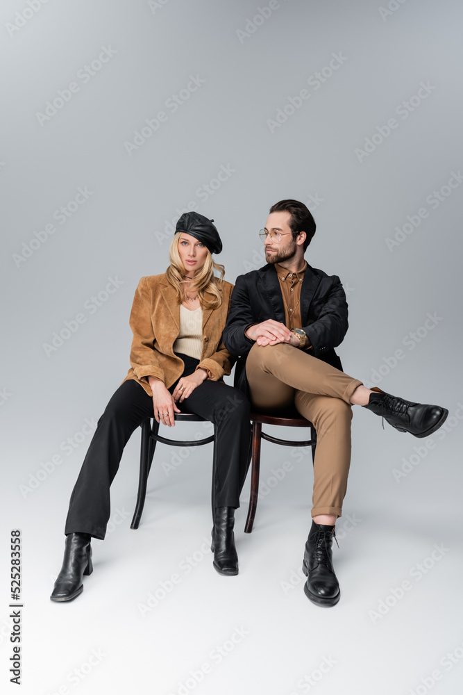 full length of blonde woman in stylish beret near bearded man in sunglasses sitting on chairs on grey full length of blonde woman in stylish beret near bearded man in sunglasses sitting on chairs on grey