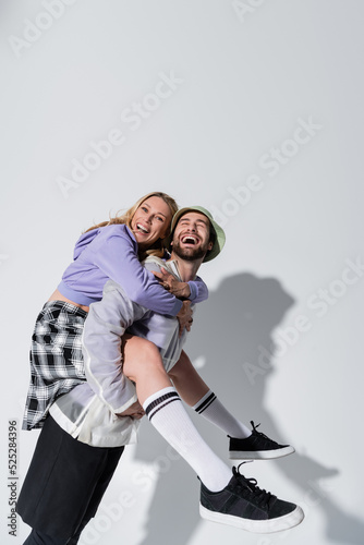 amazed man in panama hat piggybacking joyful woman in longs socks and sneakers on grey
