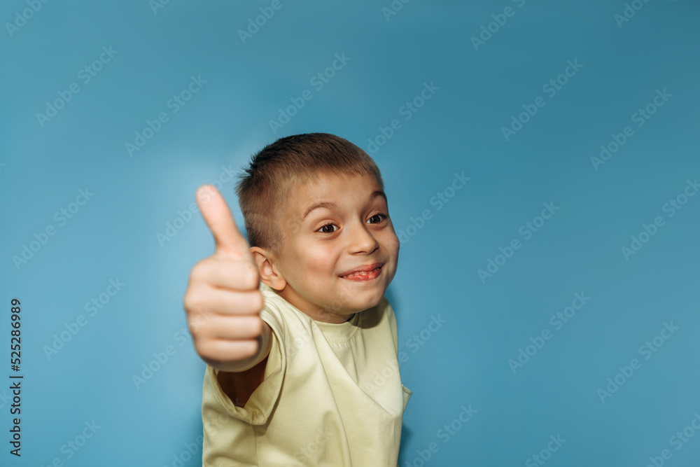 A joyful boy of 6-7 years old in a yellow t-shirt on a blue background close-up shows thumbs up. Front view A joyful boy of 6-7 years old in a yellow t-shirt on a blue background close-up shows thumbs up. Front view
