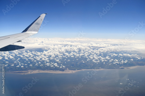 See-through aircraft window with a wing flying on a beautiful view of horizon blue sky white clouds floating over the sea, beaches meet a green land on a sunny day.