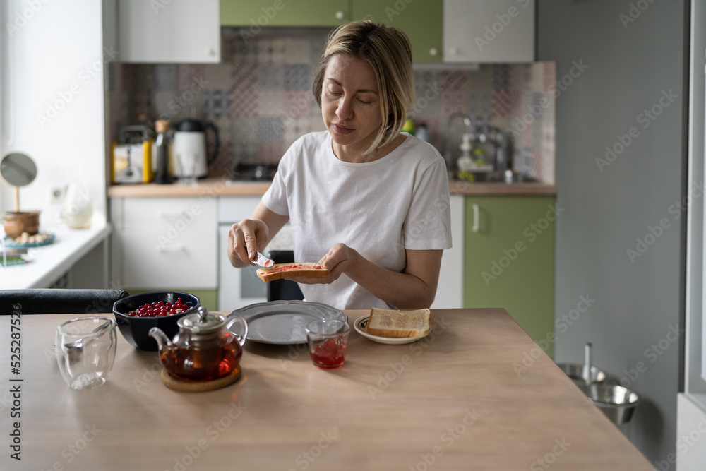 Middle-aged unkempt woman spreads jam on toast having breakfast alone. Mature blonde female eats healthy food in morning thinking about own lifestyle. Lady prepares humble breakfast in late morning