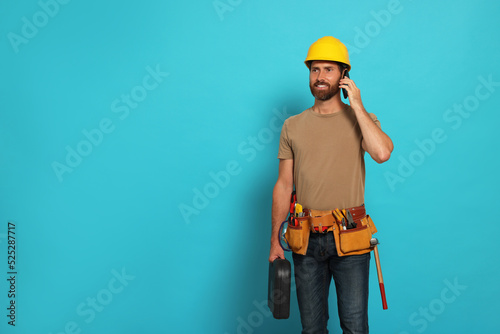 Professional builder in hard hat with tool belt talking on phone against light blue background, space for text © New Africa