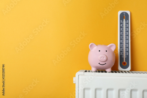 Piggy bank and thermometer on heating radiator against orange background, space for text