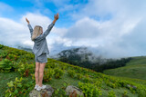 Young slender blonde woman in shorts and a shirt stands on a rock with her hands raised up on a mountain in the clouds. Bakhmaro, Georgia. Concept of travel, freedom, enjoyment