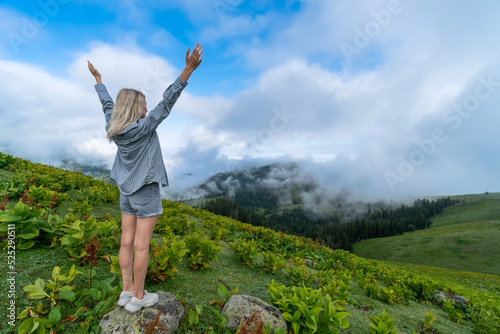 Young slender blonde woman in shorts and a shirt stands on a rock with her hands raised up on a mountain in the clouds. Bakhmaro, Georgia. Concept of travel, freedom, enjoyment