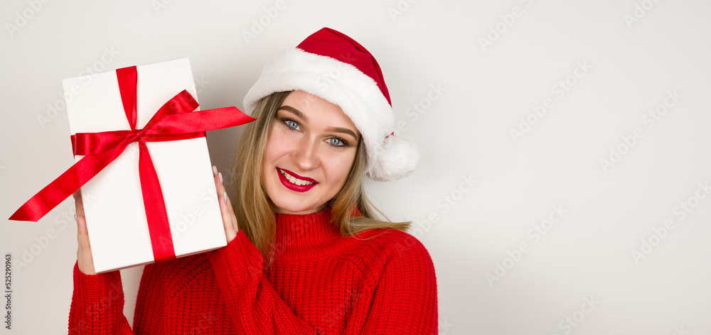 Closeup portrait of blonde girl wearing santa hat,red cozy sweater and holding gift in her hands.Banner with place for text Closeup portrait of blonde girl wearing santa hat,red cozy sweater and holding gift in her hands.Banner with place for text