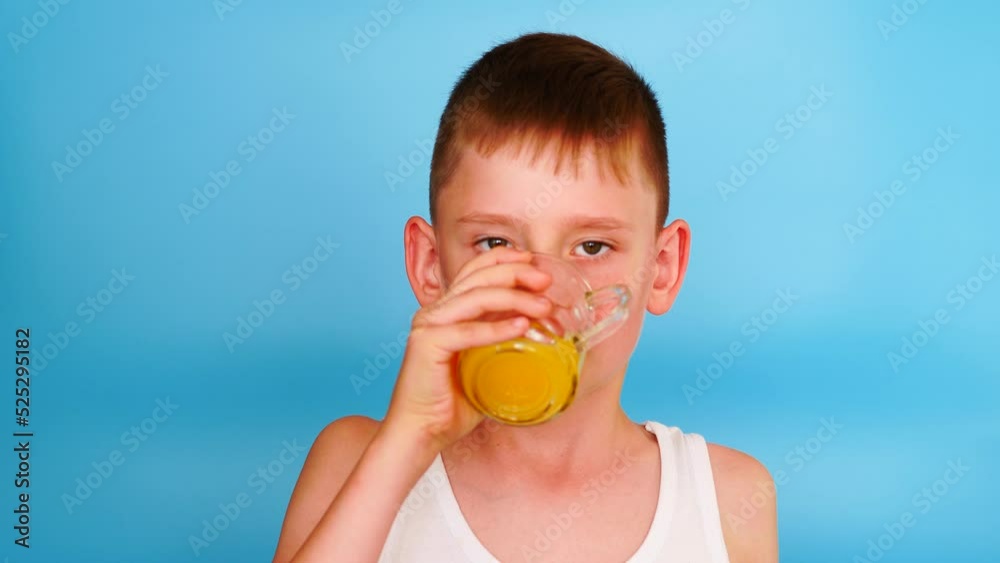 cute smiling caucasian boy 7-8 years old in a t-shirt looking at the camera drinking orange juice from a glass cup. studio shot on a blue background. the child drinks juice for breakfast cute smiling caucasian boy 7-8 years old in a t-shirt looking at the camera drinking orange juice from a glass cup. studio shot on a blue background. the child drinks juice for breakfast