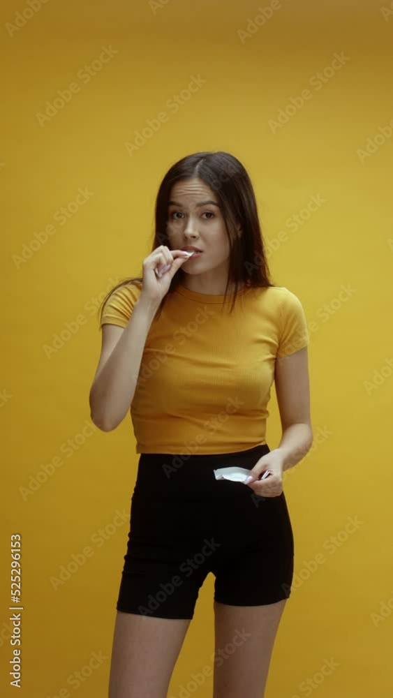 A young Asian woman putting chewing gum into her mouth, in front of a yellow background