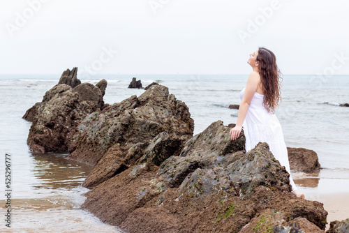 young brunette woman leaning on a rock on the beach in a white dress