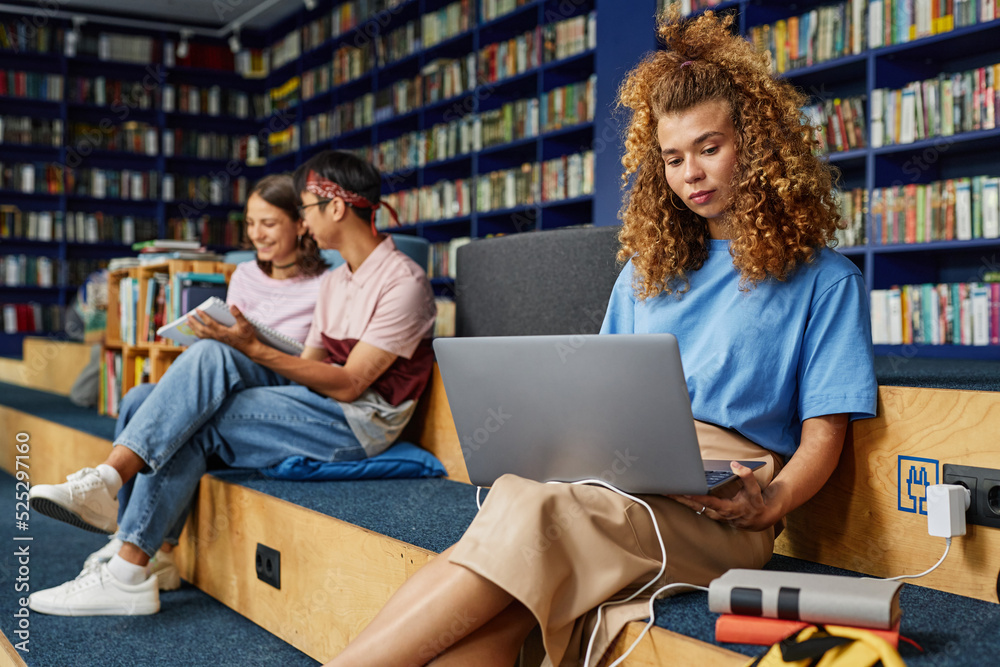 Portrait of curly haired young woman using laptop in college library and studying with people in background Portrait of curly haired young woman using laptop in college library and studying with people in background