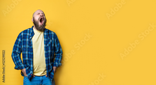Bearded middle-aged bored yawn man over yellow background. Mock up, template, copy space. Brutal, handsome. Casual clothing, plaid blue shirt, jeans.