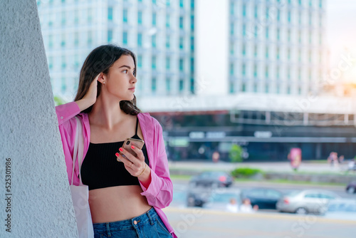 Pretty teen girl with phone in hands standing at city street outside side view