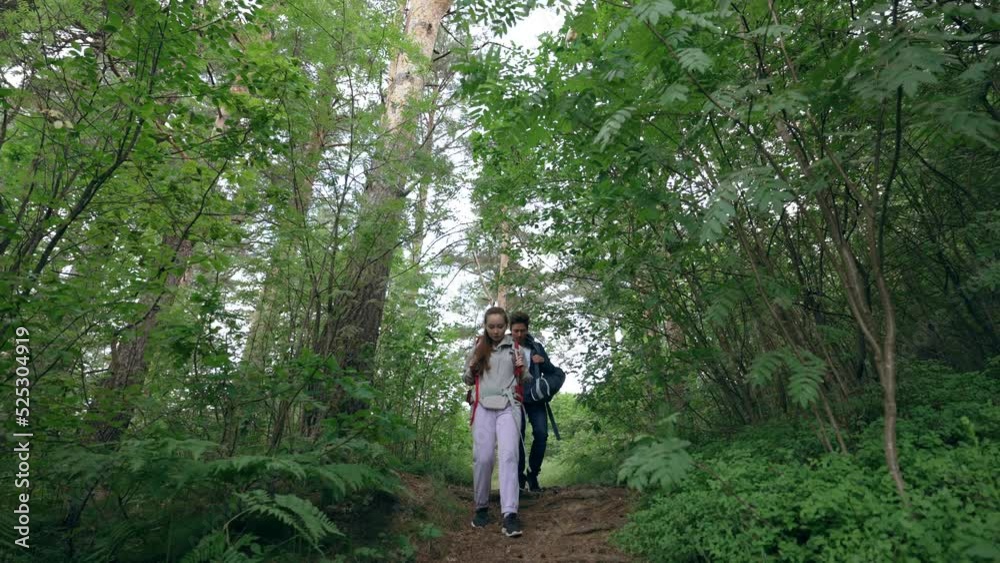 Couple of young tourists discovers various locations in deep dark forest together. Man and woman hikers with backpacks walk among thick trees slow motion Couple of young tourists discovers various locations in deep dark forest together. Man and woman hikers with backpacks walk among thick trees slow motion