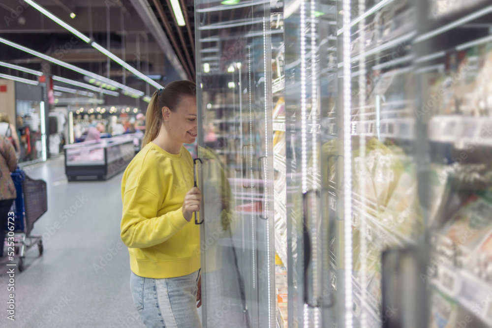 Fototapeta premium a woman of European appearance with blond hair tied in a ponytail in a yellow sweater and jeans, in a store in a refrigerated display case chooses products for the home. The concept of home food