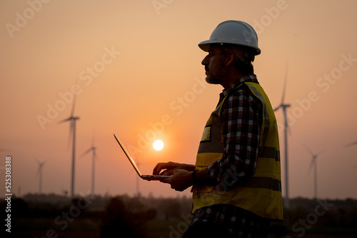 Portrait of engineer wearing yellow vest and white helmet using a computer laptop on site at wind turbines field or farm, Sustainable energy concept