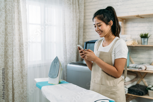 side portrait cheerful asian housewife standing by an ironing board is texting messages on the phone in a modern laundry room at home with daylight.