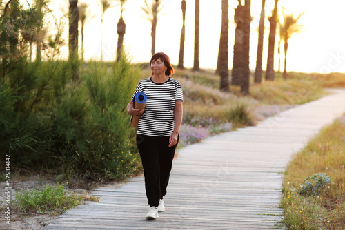 Active middle ages woman with yoga mat and walking to beach to practice yoga, exercising outdoors with happiness feeling on sunrise in the morning. Concept of healthy lifestyle, wellbeing and fitness. photo