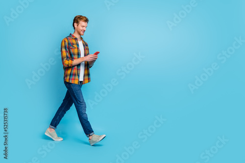 Full size photo of handsome stylish guy red hairstyle dressed checkered shirt look in smartphone walk isolated on blue color background © deagreez