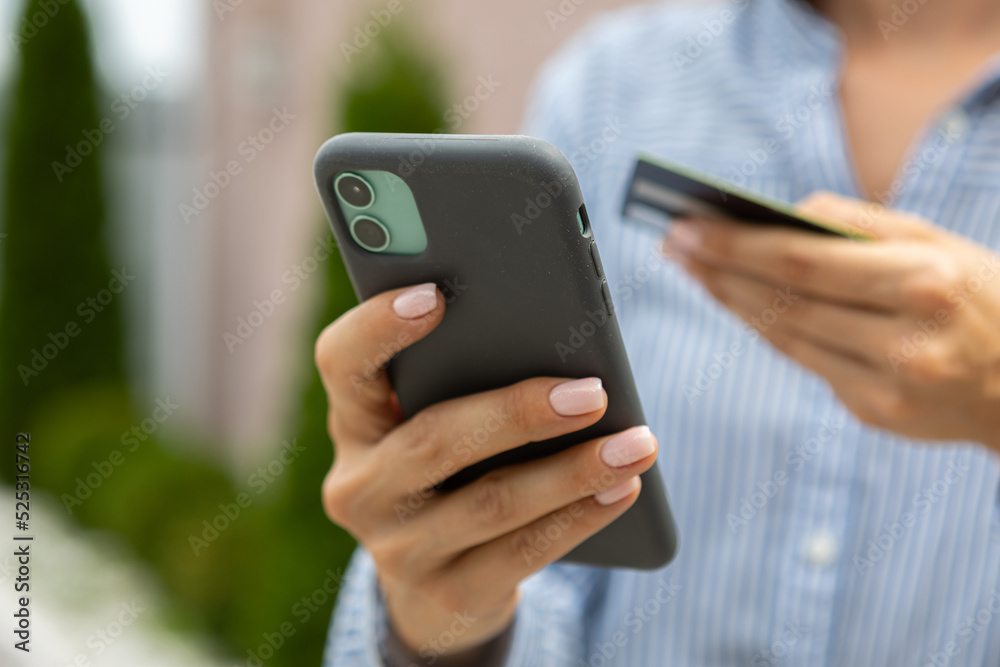 female hands hold a modern mobile phone and a bank credit or debit card on the street..ordering purchases, working online, transferring money by phone female hands hold a modern mobile phone and a bank credit or debit card on the street..ordering purchases, working online, transferring money by phone
