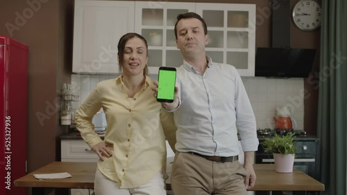 Happy couple using smartphone at dining table in kitchen,checking bank accounts,shopping online,checking social networks, ordering food from restaurant. They show the green screen phone to the camera. photo