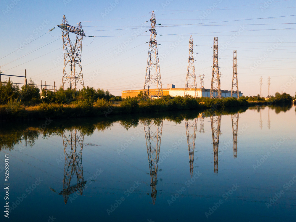 A row of metallic transmission towers of various shapes reflecting in the still water of a canal against blue sky at dusk.