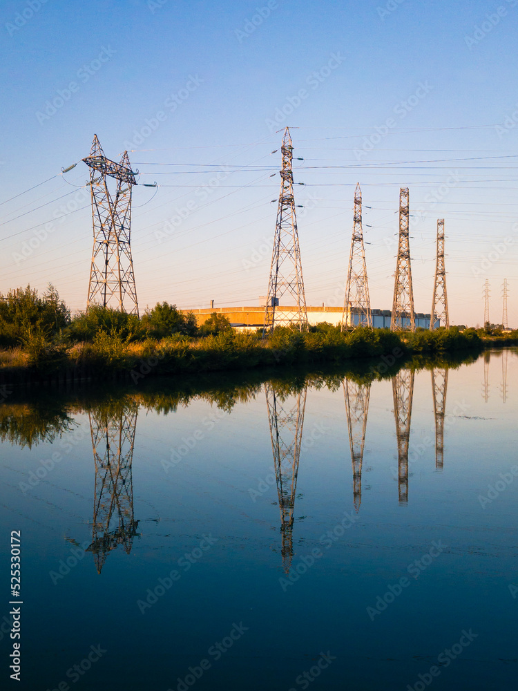 A row of metallic transmission towers of various shapes reflecting in the still water of a canal against blue sky at dusk.