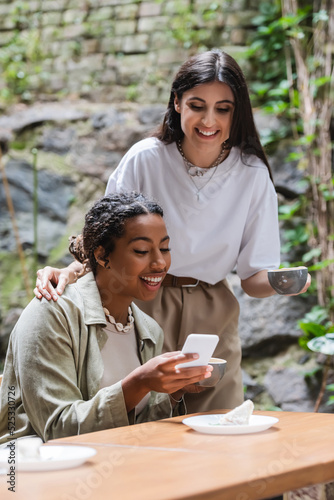 Smiling woman holding cup near african american friend with smartphone in outdoor cafe