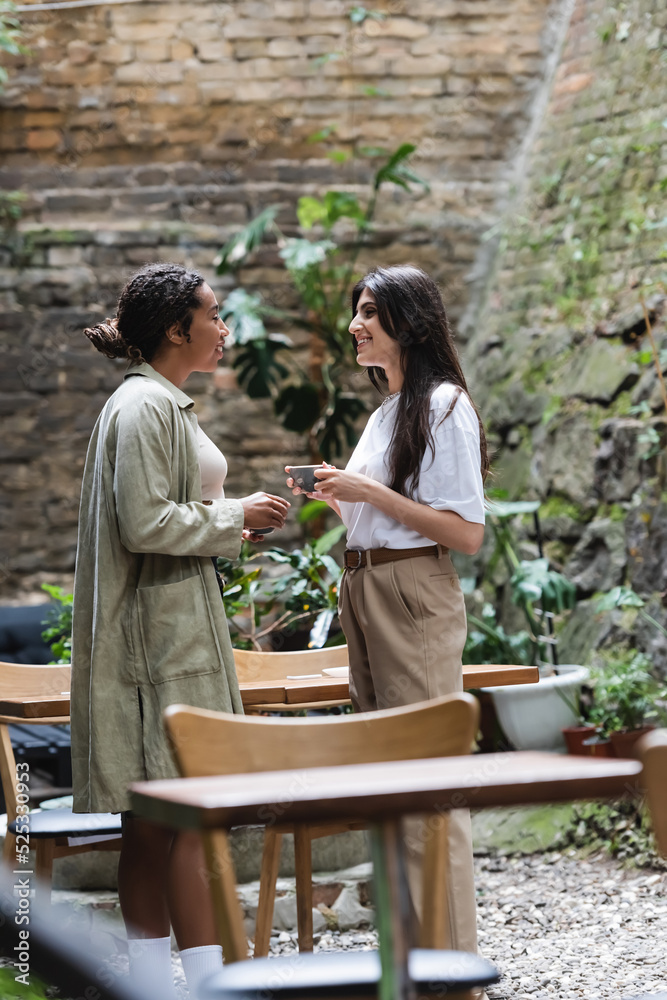Side view of multiethnic girlfriends holding coffee cups while talking on terrace of cafe Side view of multiethnic girlfriends holding coffee cups while talking on terrace of cafe