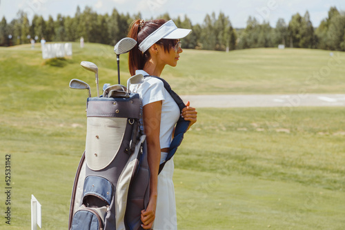 Young woman golfer in white carrying a golf bag and looking towards