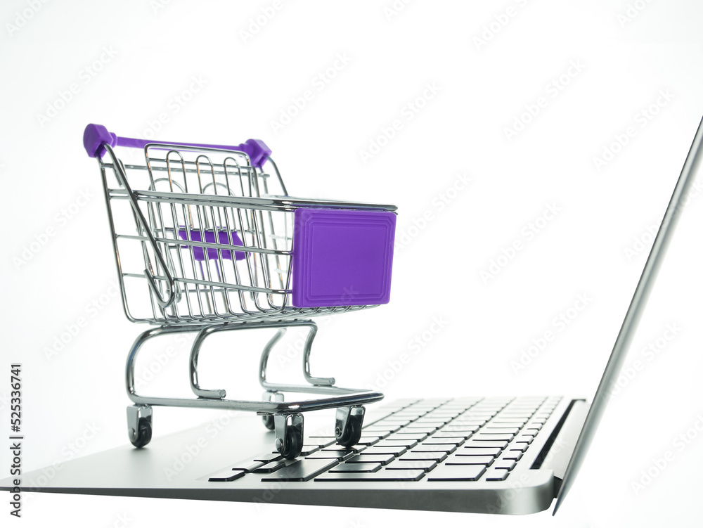 Laptop with small shopping cart on the white background, side view, close up. Toy grocery cart and notebook. The concept of electronic purchases, e-commerce.