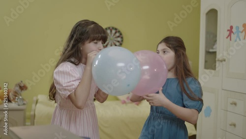 Little girls are blowing air into the balloon. Happy birthday party, celebration. Children are having fun by inflating balloons in their rooms. Cute kids inflate the balloon for their birthday.