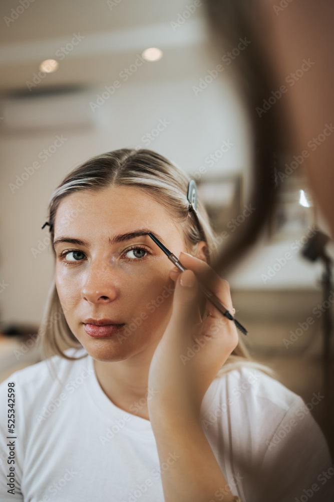 Close up shot of beautiful young woman face getting make-up. The artist is applying eyeshadow on her eyebrow with brush. Professional beautician work.