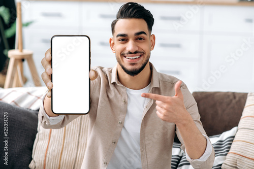 Phone screen mockup. Friendly smiling indian or arabian guy, showing smart phone with empty white mock-up screen, copy-space for advertising or presentation, sits on a sofa in a living room interior photo