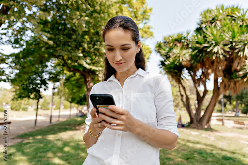 Obraz na płótnie Adorable lobby woman with long dar hair in whit shirt walking in summer green park with smartphone. European woman is using phone while standing in the park. w salonie