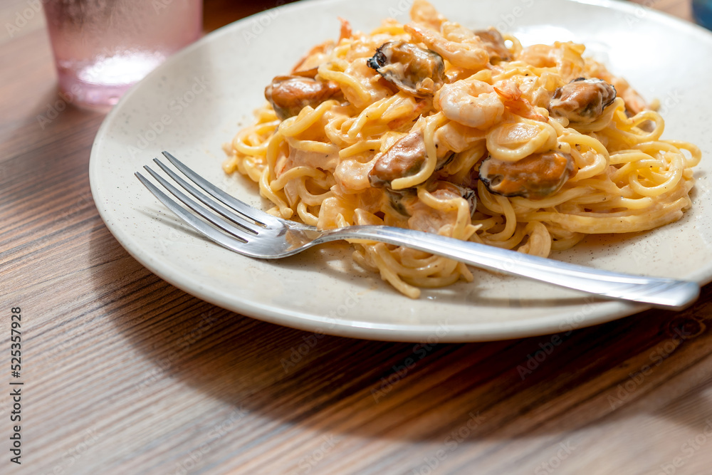 A white plate on a wooden table with pasta with mussels and shrimp, a fork and a glass of water.