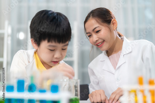 Pretty woman reacher looking at young Asia kid do experiment in laboratory at school. Smart boy use dropper to transfer colorful solution into beaker or flask. Chemical and learning elementary class