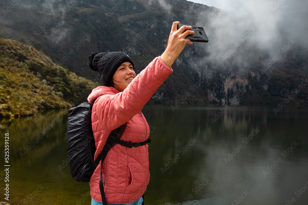 Chica con una chaqueta rosa con una mochila parada junto al lago en las montañas y tomando selfies - obrazy, fototapety, plakaty 