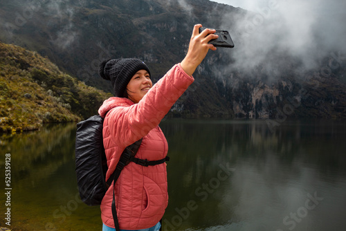 Chica con una chaqueta rosa con una mochila parada junto al lago en las montañas y tomando selfies