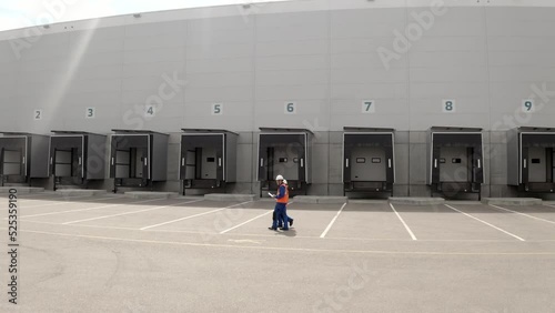 Engineers in orange vests walk against large warehouse with loading docks and parking zone. Man and woman inspect territory of logistics center aerial view