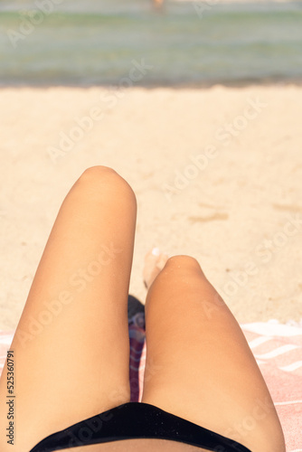 Detail of the legs of an unrecognizable woman sunbathing on the beach in a black bikini with the sea in the background.